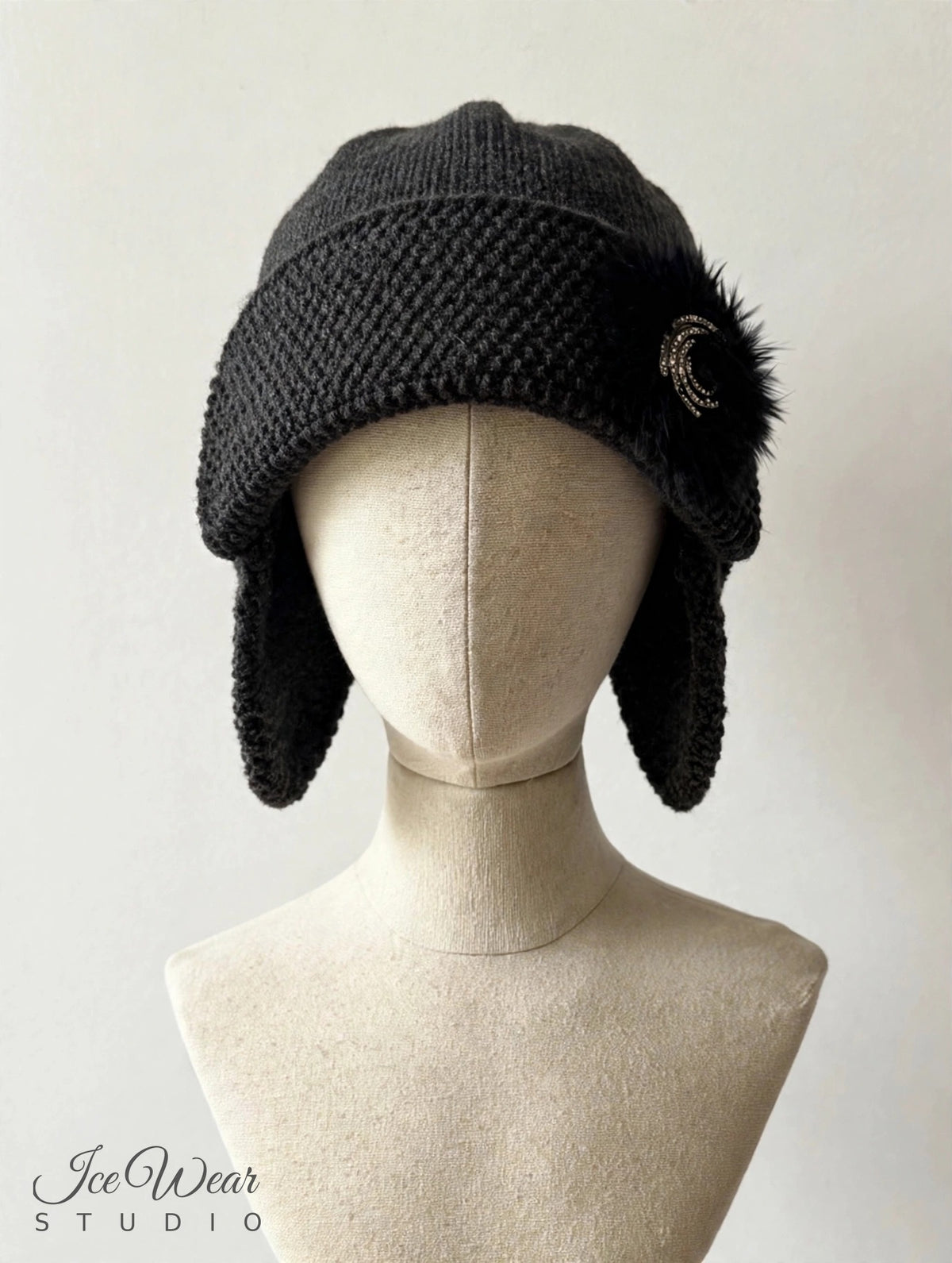 Black hand-knitted earflap cloche with fur pom and rhinestone crescent brooch, front view on linen mannequin